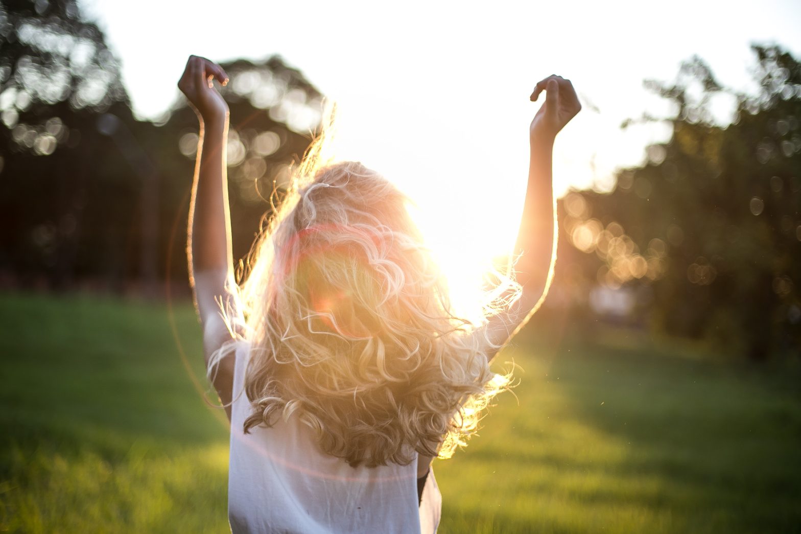 Achteraanzicht tegen de zon in, buiten in de natuur, met een blonde, langharige vrouw die haar handen omhoog houdt.